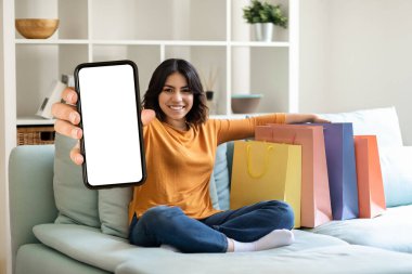 Alışveriş Uygulaması. Neşeli genç Arap kadın kamerada boş akıllı telefon gösterisi yapıyor alışveriş poşetlerinin yanında koltukta oturuyor, gülümseyen Orta Doğulu Kadın Cep Telefonu Uygulaması, Mobil Mobil Uygulama