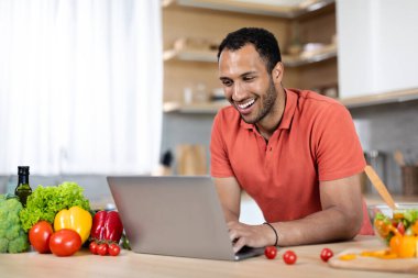 Kırmızı tişörtlü, gülümseyen, yakışıklı, bilgisayarda yazan, mutfakta organik sebzelerle sohbet eden bir adam. Yeni normal, evde iş ve iş, yemek blogu ve teknoloji