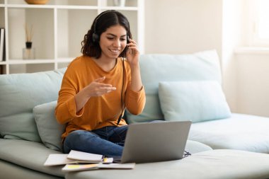 Çevrimiçi Öğretmenlik. Kulaklıklı gülümseyen Arap kadın dizüstü bilgisayarla telekonferans yapıyor, Orta Doğulu genç bayan sanal ders sırasında web kamerasında konuşuyor ve el kol hareketleri yapıyor, evdeki koltukta oturuyor.