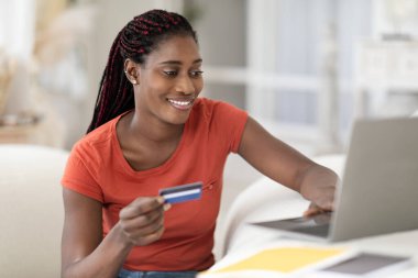 Çevrimiçi ödemeler. İçişleri Bakanlığı 'nda masasında otururken dizüstü bilgisayar ve kredi kartı kullanan gülümseyen siyah kadın, internetten alışveriş yapan neşeli Afrikalı Amerikalı kadın, e-ticaretin tadını çıkartıyor, bedava alan arıyor.