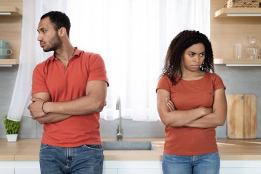 Sad offended young black man in red t-shirt ignores woman after quarrel, think about breakup at kitchen interior. Relationship problems, stress, scandal and emotions at home during covid-19 outbreak clipart