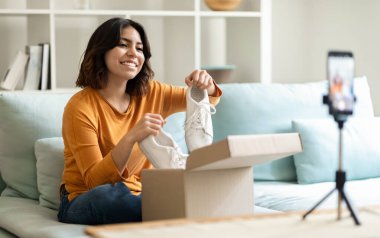 Moda videosu için boks videolarını açan gülümseyen Orta Doğulu kadın, evdeki kameranın önünde yeni spor ayakkabıları olan mutlu, genç Arap kadın paketini açıyor, üç ayaklı sandalyede akıllı telefon kullanıyor.