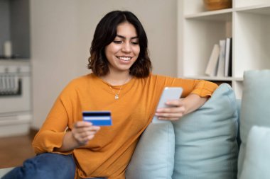 İnternetten alışveriş. Evdeki oturma odasında koltukta dinlenirken akıllı telefon ve kredi kartı kullanan gülümseyen genç Arap kadın, güzel Orta Doğulu kadın mobil ödemeler veya internet alışverişleri yapıyor.