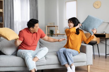 Orta yaşlı deli bir adam ve ev elbiseli genç bir kadın kanepede oturuyor, yastık savaşı yapıyor ve gülüyor, mutlu Çinli çift, evde eşleri birlikte eğleniyor, boşluğu kopyalıyor.