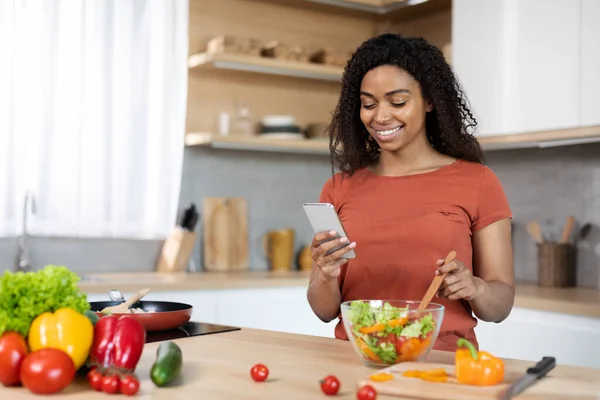 Happy young black lady in red t-shirt prepares food with organic vegetables, types on phone, reads message in kitchen interior. Cook salad for family, vegan, new app, food blog, social media at home
