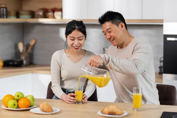 stock image Loving asian couple having conversation while eating at kitchen, cheerful loving husband taking care of his wife, pouring fresh orange juice, eating fruits and pastry, enjoying time together