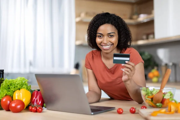 Smiling young black woman in red t-shirt with computer show credit card at table with vegetables in modern kitchen interior. Online shopping, sale and pay for goods order, business at home and finance