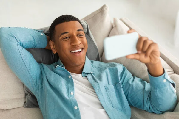 Mutlu Milenyum Afrikalı Amerikalı adam, oturma odasında boş zamanlarında akıllı telefondan komik videolar izleyip, görüntülü görüşme yapıyor. Cihazla evde dinlen ve rahatla, covid-19 karantinası sırasında yeni uygulama