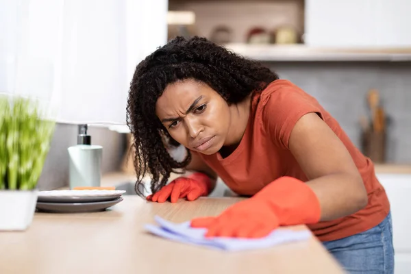 Dissatisfied sad young black lady in red t-shirt, rubber gloves wipes dirt from wooden table in kitchen interior. Housework, perfectionism, problems with dust cleaning, household chores, perfectionism