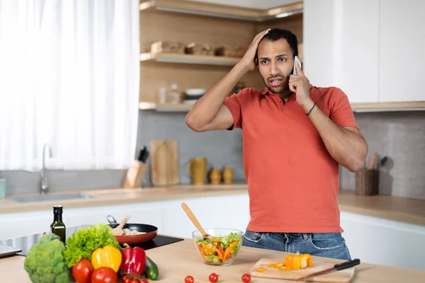 Sad confused young african american guy in red t-shirt holds his head with hand calls by phone, got bad disturbing news on kitchen interior, copy space. Problems at home, stress and people emotions