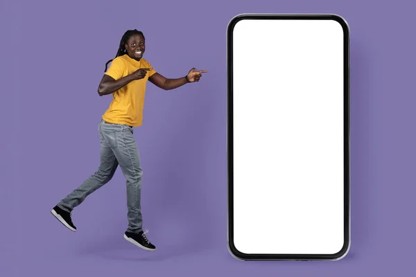 用巨大的智能手机随意跳着长长的辫子 指着白色的空白屏幕 展示最新的移动应用程序 模型和紫色工作室背景的富有感情的 时尚的黑人小伙子 — 图库照片
