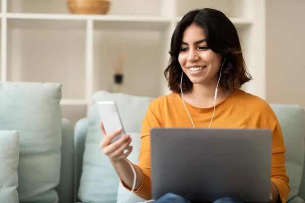 数码娱乐 带着笔记本电脑和智能手机在家里休息的中东女性的微笑 带着耳机坐在客厅的长椅上 使用现代家用电器的快乐阿拉伯女性 — 图库照片