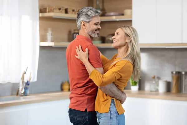 Happily married middle aged couple handsome grey-haired man and blonde woman dancing at kitchen, loving spouses having romantic time at home, wearing casual comfy outfits, copy space