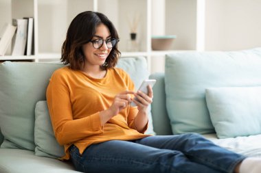 Genç Arap kadın gözlük takıyor evde akıllı telefonuyla rahatlıyor, koltuğa oturmuş gülümseyen Orta Doğulu kadın portresi ve online alışveriş veya iletişim için cep telefonu kullanıyor.