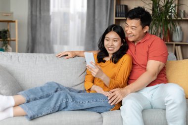 Neşeli Asyalı çift evde birlikte vakit geçiriyorlar, pozitif orta yaşlı erkek ve genç kadın oturma odasında dinleniyorlar, akıllı telefon kullanıyorlar, mobil oyun oynuyorlar, web sörfü yapıyorlar, uzay kopyaları kopyalıyorlar.