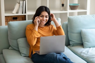 Neşeli genç Arap kadın cep telefonuyla konuşuyor ve evinde dizüstü bilgisayar kullanıyor, bilgisayar ekranına bakıp gülümsüyor ve keyifli cep telefonu konuşmaları yapıyor.