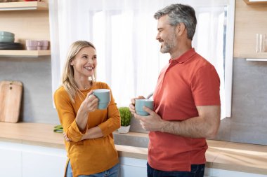 Neşeli, yakışıklı, gri saçlı orta yaşlı adam ve güzel sarışın kadın evde çay içerken, kahve fincanları tutarken, birbirinize bakarken, gülümserken, mutfak içindeyken.