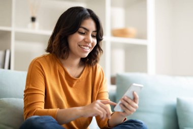 Evde dinlenirken akıllı telefondaki mutlu genç Arap kadın mesajı, koltukta dinlenen ve çevrimiçi iletişim için cep telefonu kullanan gülümseyen Orta Doğulu kadının portresi,