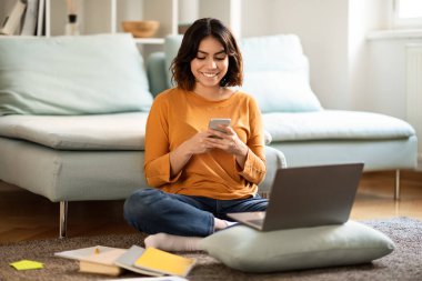 Evde dizüstü bilgisayarı ve akıllı telefonuyla dinlenen mutlu genç Arap kadın, odada oturmuş cep telefonuyla mesajlaşan neşeli Orta Doğulu kız öğrenci, çevrimiçi iletişimin keyfini çıkarıyor.