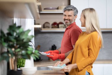 Yakışıklı, orta yaşlı, kır saçlı koca karısına ev işlerinde yardım ediyor yemekten sonra bulaşıkları yıkıyor, sohbet ediyor ve gülümsüyor.