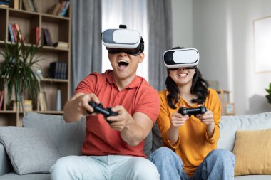 Mutlu Koreli çift evde video oyunu oynuyor, neşeli orta yaşlı adam ve güzel genç kadın kanepede oturuyor, ellerinde joystickler ve sanal gerçeklik gözlüğü kullanıyorlar, kopyalama alanı. Ev eğlencesi konsepti