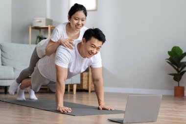 Aktif Koreli eşler evde yan yana yatıyorlar, pozitif Asyalı orta yaşlı erkek ve genç kadın yoga minderi üzerinde egzersiz yapıyorlar, kadın kocasına sırt üstü uzanıyor, modern dizüstü bilgisayara bakıyor, uzayı kopyalıyor.