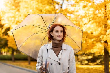 Sonbaharda şehir parkında sarı yapraklarla yağmurda şemsiyeli, kızıl saçlı, ciddi, Avrupalı genç bir bayan yalnız yürür. Hafta sonları boş zaman, kötü havada duygular, covid-19.