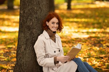 Gülümseyen Avrupalı kızıl saçlı güzel bayan sonbaharda şehirde sarı yapraklarla parktaki ağacın yanında kitap okuyor. Edebiyat, boş zamanlarında hobi, kadın hafta sonu yalnız başına dışarıda eğleniyor, covid-19.