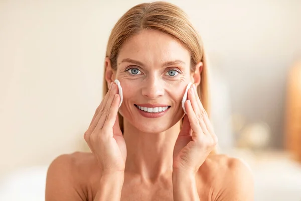 Portrait of positive middle aged woman using cotton pads, erasing make up from her face, applying facial toner or cleansing milk, bathroom interior, copy space