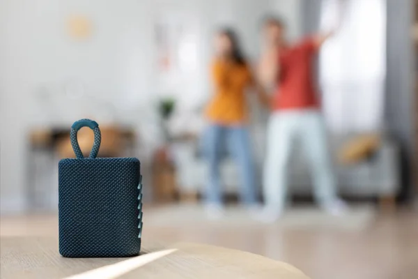 Selective focus on brand new portable smart speaker over happy singing asian couple, blurred background, copy space, home interior. Modern technologies and domestic entertainment concept
