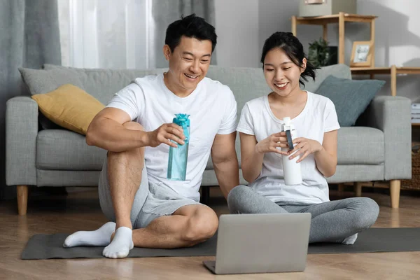 快乐的中国中年男子和年轻女子穿着运动服坐在健身垫上 在家锻炼后喝水 看着电脑屏幕笑着 复制空间 — 图库照片