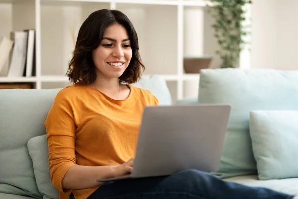 自由概念 在家里用笔记本电脑工作的年轻阿拉伯女性微笑 在客厅里坐在沙发上 用电脑远程办公的快乐的中东自由职业女性 自由空间 — 图库照片