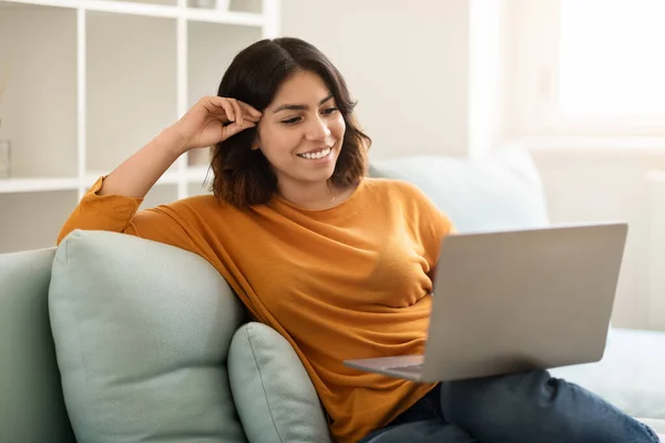 快乐的中东年轻女性坐在沙发上 用笔记本电脑 笑着坐在沙发上 看电脑屏幕 看电影 或浏览社交网站 — 图库照片