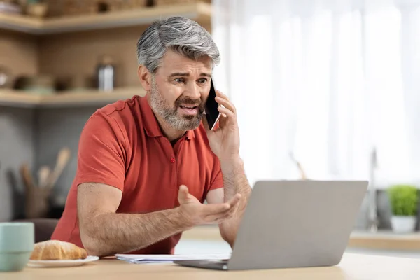 Angry handsome middle aged entrepreneur working from home, man sitting at table in front of modern laptop with cup of coffee, having phone call, looking at computer screen and gesturing