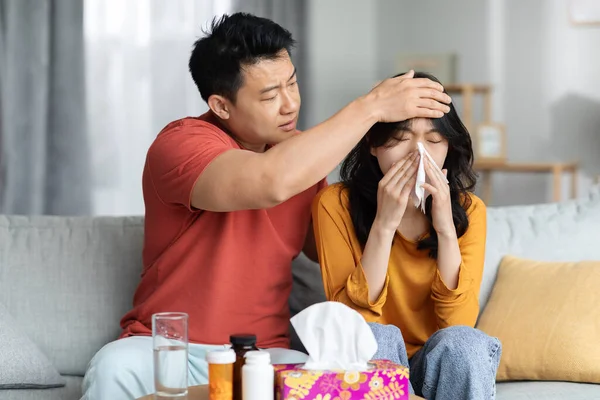 爱亚洲中年男人的丈夫照顾他生病的妻子 摸她的额头 检查发烧 生病的女人打喷嚏 用餐巾 夫妻坐在沙发上 复制空间 — 图库照片