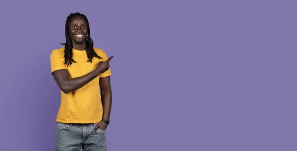 一位有着长长的辫子 穿着黄色T恤和牛仔裤的英俊的非洲裔美国年轻人指着广告在紫色工作室背景 全景的复制空间 — 图库照片
