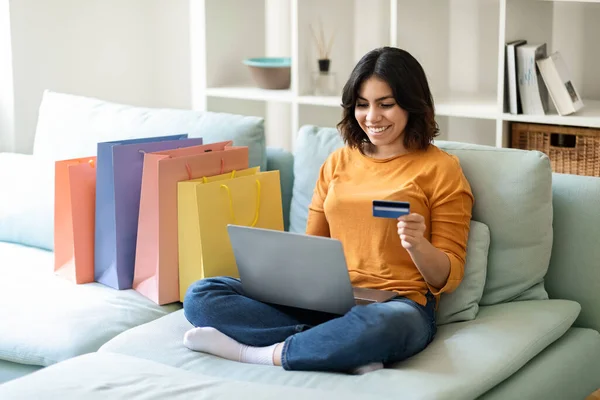 Online Shopping Concept. Smiling Arab Woman Using Laptop And Credit Card While Sitting On Couch With Bright Shopper Bags At Home, Happy Middle Eastern Female Making Pucrhases In Internet, Free Space