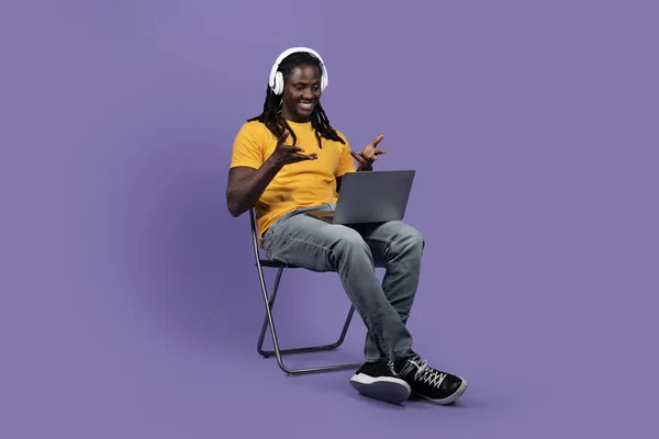 Cheerful stylish long-haired young black man sitting on chair, using brand new computer and wireless headset, having video chat with colleagues or friends, purple studio background, copy space