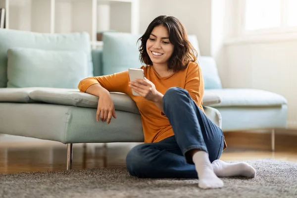 用智能手机在地板上放松一下 快乐美丽的中东女性在手机上浏览互联网 或在客厅和自由空间在线购物 — 图库照片