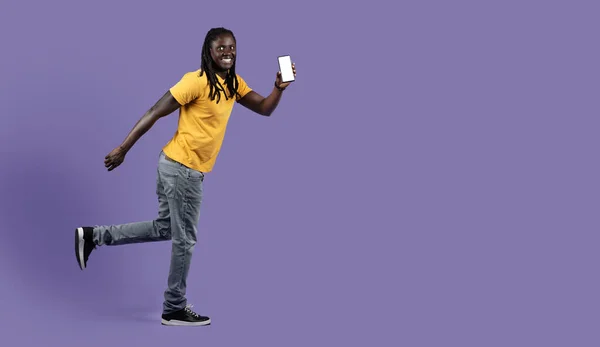 コピースペース 紫色のスタジオの背景に向かって実行している彼の手の中にスマートフォンを持つ刺激的なスタイリッシュな長髪の若いアフリカ系アメリカ人男性 白い空の画面と笑顔 モックアップ付き携帯電話を示しています — ストック写真