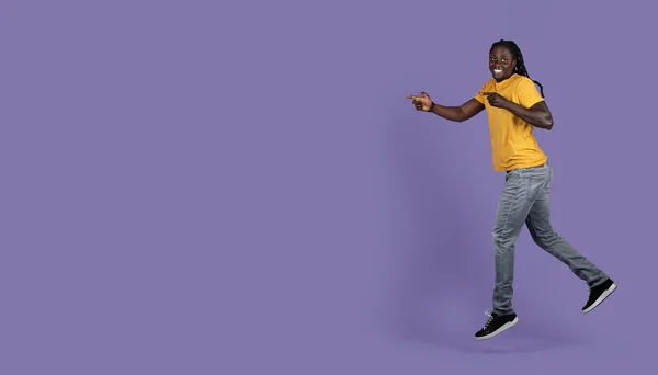 兴奋而又长发的年轻黑人小伙子在紫色的工作室背景上奔向复制的空间 摆出瞄准的姿态 指着广告 — 图库照片