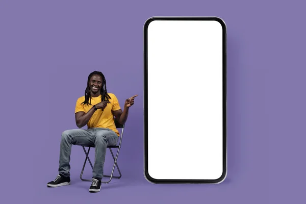 年轻的非洲千禧年男子 头戴凉爽的长辫子 穿着休闲装 坐在椅子上 坐在巨大的智能手机旁 用紫色背景的白屏指著 工作室拍摄 — 图库照片