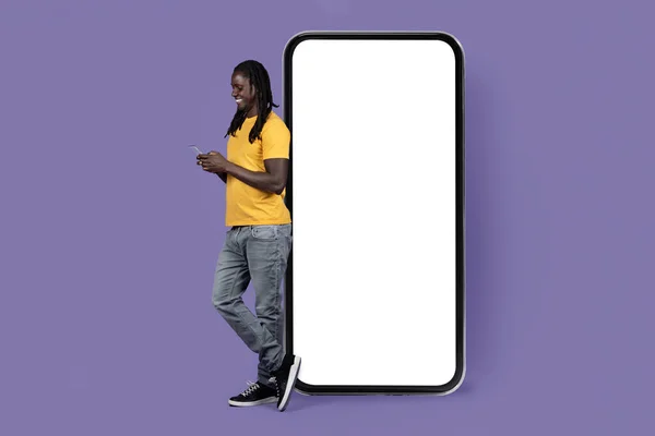 快乐的非洲裔美国千禧年男子 头戴凉爽的长辫子 穿着休闲装 站在巨大的智能手机旁边 有着紫色背景的白色空白屏幕 他使用手机 工作室镜头 — 图库照片