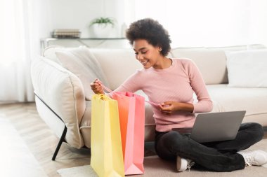Gülümseyen bin yıllık Afro-Amerikan kadın oturma odasının iç kısmında dizüstü bilgisayarla yerde oturan birçok pakete bakıyor. İyi satış, moda blogu, sipariş ve teslimat
