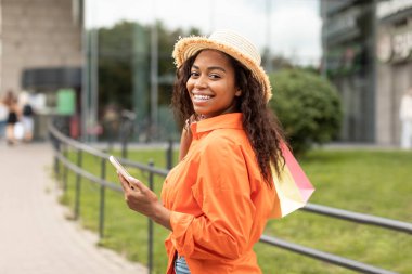 Bir sürü alışveriş çantası olan mutlu genç zenci kadın telefonda konuşuyor, şehirde yürüyüş ve satışın keyfini çıkarıyor, kameraya bakın. Gadget, sosyal mesafe, online alışveriş için yeni uygulama, bankacılık ve nakit para