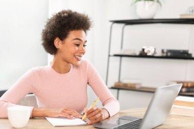 Gülümseyen Afro-Amerikan bayan not defterine notlar alıyor, ofisin içinde video bağlantısı var, fotokopi odası. Öğretmenlik, öğrenim, iş, iş ve evde uzaktan ve sosyal mesafede buluşma