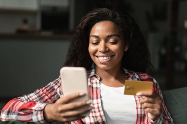 Kredi kartı ve akıllı telefon tutan, blog okuyan, oturma odasında online alışveriş yapan neşeli, güzel siyah bir kadın. Yeni uygulama, bankacılık, finans ve satın almaktan gelen nakit para, evde büyük indirim