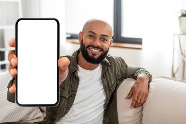 Moda cep telefonu. Elinde beyaz beyaz ekranlı akıllı telefon tutan mutlu Latin adam, kameraya yakın cihazlar gösteriyor, evdeki kanepede oturuyor. Boş boşluklu Gadget, düzenle