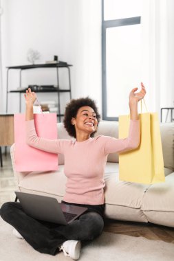 Bin yıllık Afro-Amerikan kadınının bir sürü alışveriş torbasıyla el kaldırması, büyük bir alışveriş ve başarı sevinci içinde oturma odasının içinde yerde oturmasına sevindim. Kazan, evde büyük indirim var.