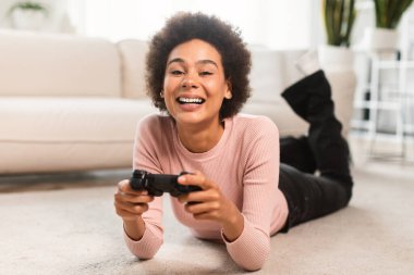 Neşeli bin yıllık Afro-Amerikan kadın elinde joystickle yerde yatıyor, oturma odasının içinde online oyunlar oynuyor, kapanıyor, mekanı kopyalıyor. Evde boş zamanlarında aygıtla eğlence